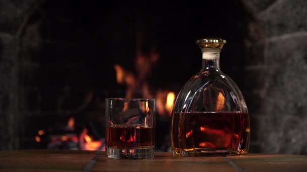 Бутылка и стакан с виски или коньяком на фоне огня в камине — стоковое видео