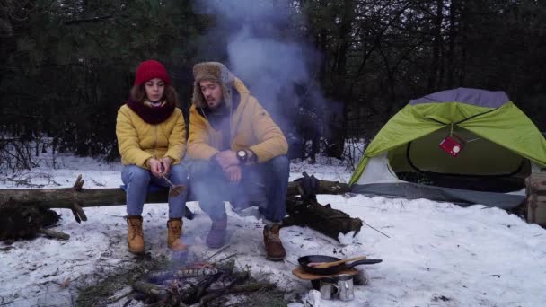 Ung man och kvinna steker korv i brand i vinterskogen — Stockvideo