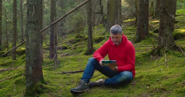 En skäggig man sitter på mossan och läser en bok. — Stockvideo