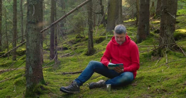 Egy szakállas ember ül a mohán és könyvet olvas. — Stock videók