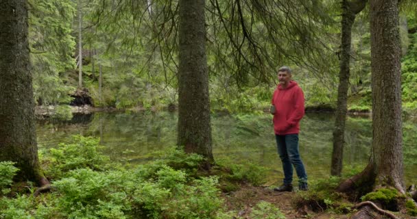 Bearded man drinks tea from a mug near a mountain lake — Vídeo de stock