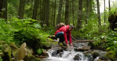 Bearded man with a backpack washes his hands with water from a mountain stream