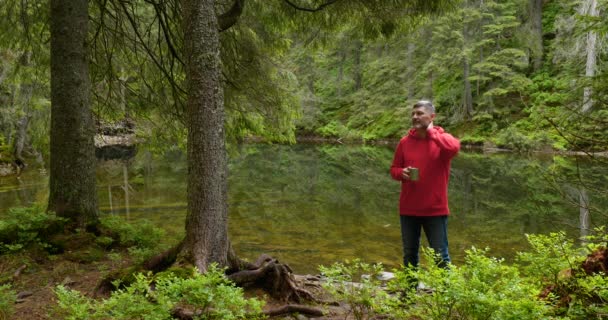 Homem barbudo bebe chá de uma caneca perto de um lago de montanha — Vídeo de Stock