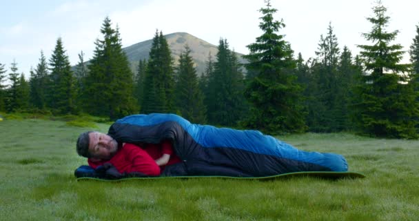 Bearded man sover i en sovsäck på en vacker äng i bergen — Stockvideo