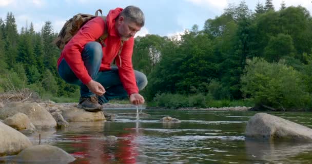 Un uomo barbuto con uno zaino beve acqua da un fiume di montagna — Video Stock