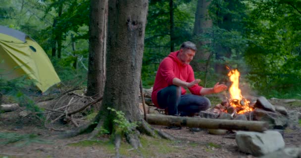 Man met baard toerist verwarmt zijn handen over een kampvuur in het bos — Stockvideo