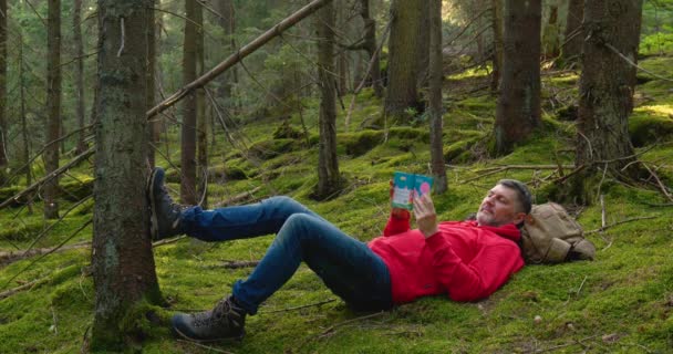 Бородатий чоловік відпочиває в лісі і читає книгу — стокове відео