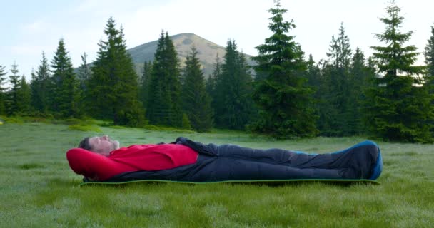 En skäggig man i en sovsäck vaknar i en skogsglänta i bergen. — Stockvideo