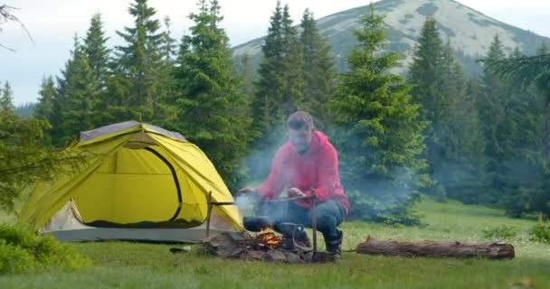 A szakállas férfi turista felmelegíti a kezét a tábortűz felett a hegyekben. — Stock videók