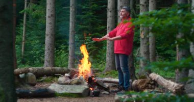 Sakallı erkek turist, ormanda bir şenlik ateşinde ellerini ısıtıyor.