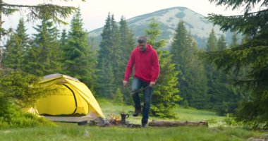 Sakallı bir adam dağlardaki güzel bir orman çimenliğinde ateş yakar.