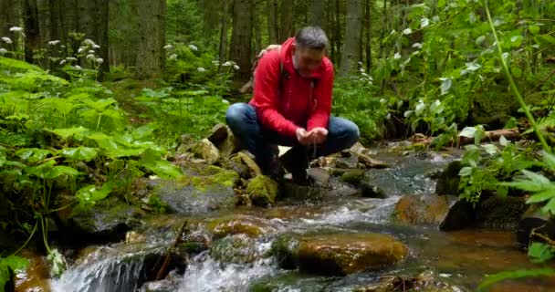 Egy szakállas férfi hátizsákkal vizet iszik egy hegyi patakból. — Stock videók