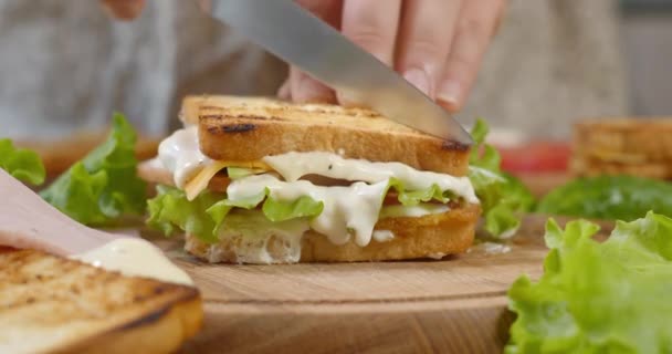 Γυναίκα σεφ κόβει κλαμπ σάντουιτς στη μέση με κοφτερό μαχαίρι σε μια ξύλινη σανίδα στην κουζίνα — Αρχείο Βίντεο