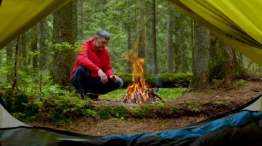 Sakallı bir adam güzel bir ormanda ateşin yanında güneşleniyor.