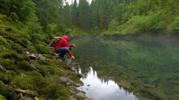 Egy szakállas férfi hátizsákkal vizet iszik egy tiszta tóból. — Stock videók