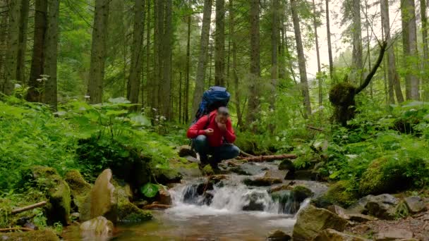 Бородач с рюкзаком пьет воду из горного ручья. — стоковое видео