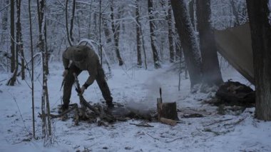 Karlı Orman Kampı 'nda yakacak odun hazırlayan bir adam, Kamp bir kış sığınağının yakınındaki dalları kesiyor ve ateş çukurundan dumanlar yükseliyor.