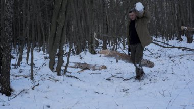 Ormanda kış çalılıkları taşıyan, haki rengi ceket ve yeşil pantolon giyen bir adam, karla kaplı bir ormanda omzunda büyük bir kütük taşıyor.
