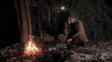 Gece kamp ateşinde sıcak içecek içen bir adam, kış ormanı barınağında, orman kampçısı, soğuk mevsimde ateşin yanında ısınıyor.