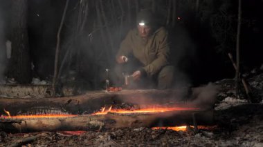 Bir adam yanan ateşin yanında, gece vakti ormandaki geçici bir barınağın altında, bir lambayla aydınlatılmış bir şekilde oturur. Bu sahne vahşi doğada hayatta kalmanın özünü anlatıyor.
