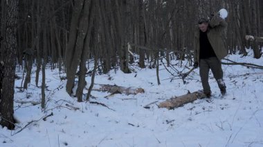 Kereste taşıyan Ormanda Kış Çalılığı, haki rengi ceket ve yeşil pantolon giyen bir adam karlı bir orman sahnesinde omzunda büyük bir kütük taşıyor.