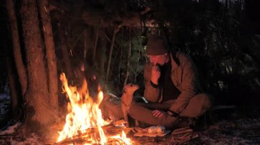 İnsan ve köpek gece orman sığınağında kamp ateşinin yanında otururken, Bushcrafter ve köpeği soğuk bir gecede ağaçlık alanda ateşin başında dinleniyorlardı.