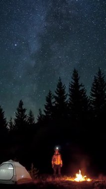 Gece karavanı ve ormanın ortasında kamp ateşi olan bir adam, kamp ateşi ve çadırın yanında duran bir adam yıldızlı gökyüzünün altında geceleyin dikey bir video.