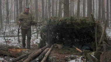 Bushcraft barınak binası ve kış ormanlarında kamp ateşi hazırlığı, açık havada hayatta kalma aktivitesi, kamp alanının yakınındaki yakacak odunları ve dalları düzenleyen bir adam.