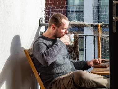 Adam güneşli bir günde balkonda kahve içiyor. 
