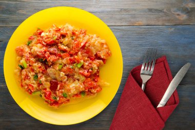 Mavi ahşap arka planda sarı tabakta soğanlı haşlanmış domates. Çatal ve bıçak manzaralı vejetaryen yemekleri.