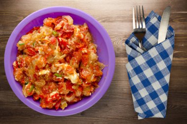 Kahverengi ahşap arka planda mor tabakta soğanlı haşlanmış domatesler. Soğan manzaralı haşlanmış domates. Çatal ve bıçakla vejetaryen yemeği