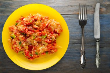 Mavi ahşap arka planda sarı tabakta soğanlı haşlanmış domates. Çatal ve bıçak manzaralı vejetaryen yemekleri.