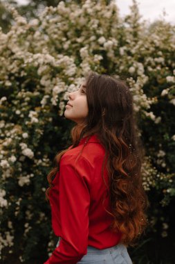 Kıvırcık kızıl saçlı güzel bir kızın çiçek açan çalıların yanında duruşu.