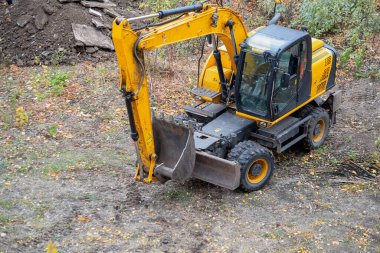 A working excavator with a visible cab and bucket on the ground among shrubs.