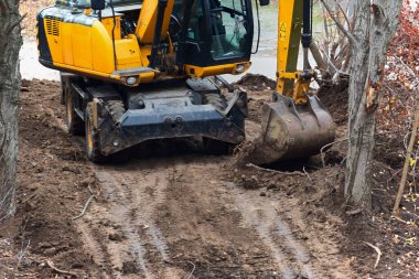 An excavator digs the ground on a site with trees, demonstrating work in confined spaces and the need for careful treatment of the environment.