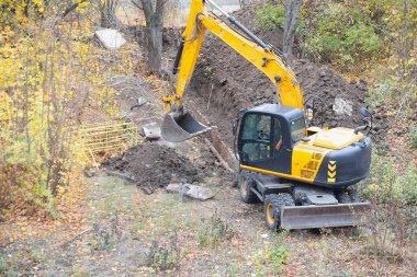 The excavator digs a deep pit on a construction site surrounded by trees.