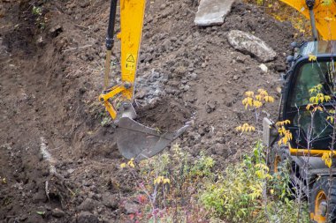 Close-up of an excavator bucket full of soil against the background of a dug trench during construction work, excavation of soil using the excavator bucket, the first stage of construction work.