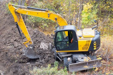 Sarı bir kazıcı, çalılarla çevrili bir alanda toprak taşıma işi yapar..