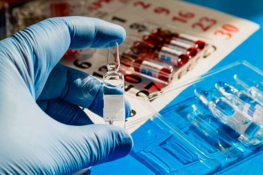 Doktorun elindeki bir ampulün tarihli takvimde yakın plan görüntüsü. Tıbbi planlamanın ve aşı takvimine uymanın bir sembolü..