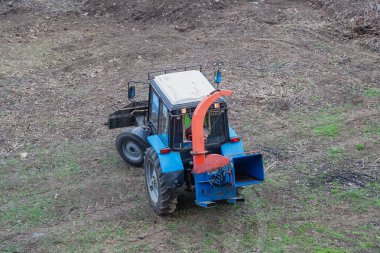 Traktörün üzerinde seyyar bir odun öğütücü parkta çalışmaya hazır bekliyor. Modern makineler, kentsel bir ortamda odun atıklarının çevre dostu bir şekilde bertaraf edilmesine yardımcı olur..