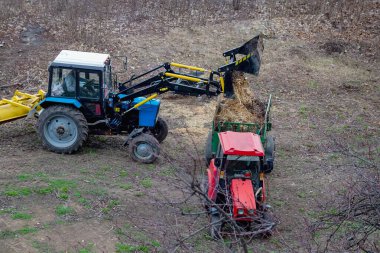 Bir inşaat yükleyicisi, bölgedeki başka bir traktörün attığı tahta atıklarını atmak için minibüse yüklüyor. Bu da araziyi çevre dostu bir şekilde düzenlediğini gösteriyor..