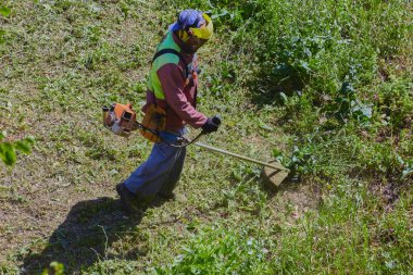Kavurucu bir yaz gününde, tulumlu bir bahçıvan ve miğfer, çim biçme makinesiyle uzun otları biçer..