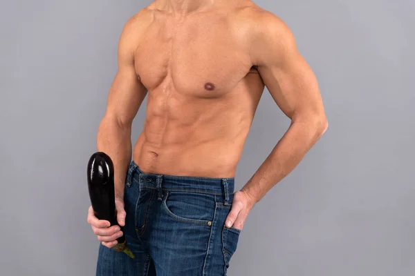 Muscular man with six pack abs torso cropped view hold firm and large eggplant at crotch level grey background, penis enlargement.