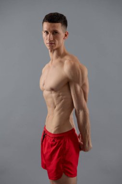 Göbek, göğüs ve pazılara sahip kaslı bir adam spor şortuyla poz veriyor gri arka plan, vücut geliştirici..