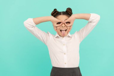 Mutlu okul çağında çocuk parmaklarını süper kahraman maskesi yapar dilini mavi arka plana sokar, oynar.