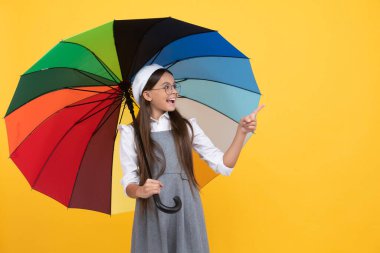 Gözlüklü mutlu liseli kız. Renkli güneş şemsiyesi altındaki genç çocuk Finegr 'i işaret ediyor. Gökkuşağı şemsiyeli çocuk. Sonbahar sezonu. Yağmurlu hava tahmini. Okula dönüyorum. Canlı yağmur koruması. Boşluğu kopyala.