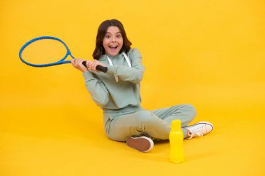 Mutlu çocuk elinde su şişesi, spor kıyafetleri, salıncak tenis raketi, sarı arka planda topa vurmak için oturur..