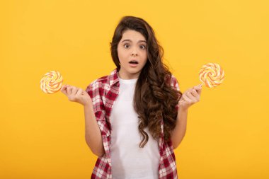 Şok olmuş kıvırcık saçlı genç kız sarı arka planda lolipop karamel şekeri ve şekerleme tutuyor..