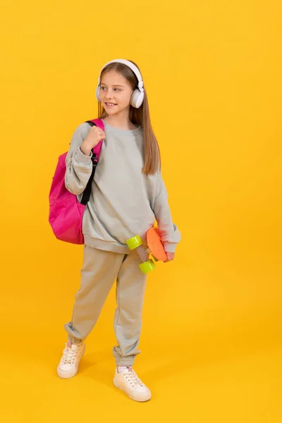 Szczęśliwy nastolatek dziewczyna dzieciak z plecakiem szkoły i penny pokładzie słuchać muzyki w słuchawkach, czas szkoły. — Zdjęcie stockowe