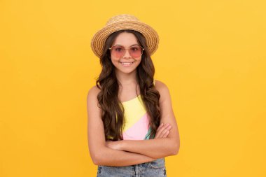 Yazın hasır şapkalı ve gözlüklü mutlu çocuk sarı arka planda kıvırcık saçlı, plaj modasında.